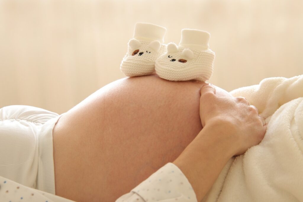 Femme enceinte posant les mains sur son ventre avec deux chaussons de bébé, illustrant l’accompagnement ostéopathique de la grossesse.